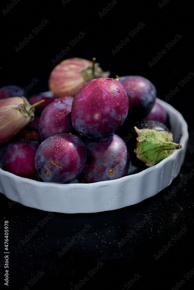 Seasonal Blue and purple Fruit and vegetable bowl with green leaves in a bowl on rustic dark background, top view. Plum and Organic Raw Baby Indian Eggplants Seasonal Blue and purple Fruit and vegetable bowl with green leaves in a bowl on rustic dark background, top view. Plum and Organic Raw Baby Indian Eggplants