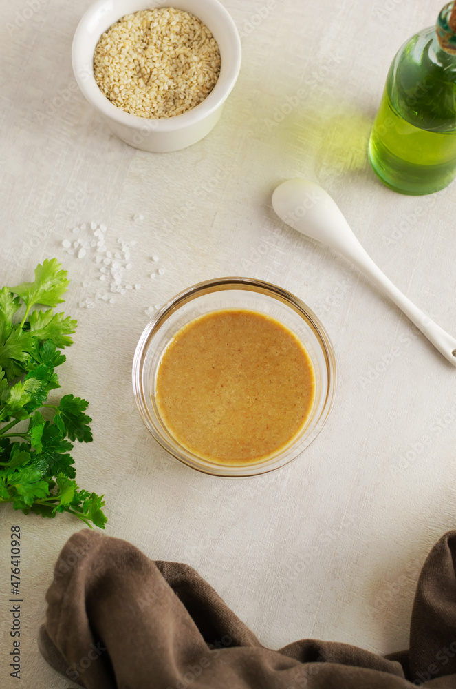 Tahini in a glass bowl with ingredients and herbs on a light background. The process of preparing a traditional Asian sauce. Levantine cuisine concept. Tahini in a glass bowl with ingredients and herbs on a light background. The process of preparing a traditional Asian sauce. Levantine cuisine concept.