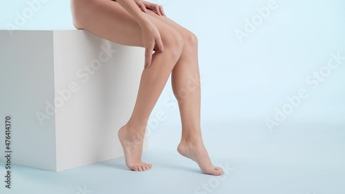 Horizontal medium shot of slim young good-looking woman in black underwear strokes her tiptoe foot and leg sitting on the white platform on pale blue background | After woman shaving concept photo