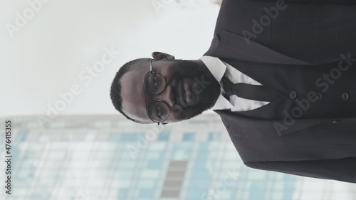 Handheld vertical portrait of happy African-American businessman in three-piece suit and glasses smiling for camera and posing in city. Building in background photo