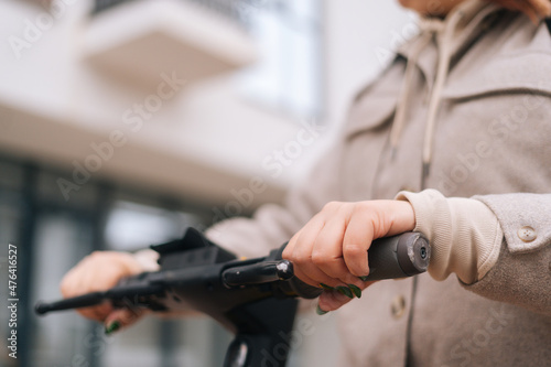 Close-up hands of unrecognizable young woman riding on electric scooter in city street on blurred background of building. Female holding steering wheel of electric scooter and ready to go.