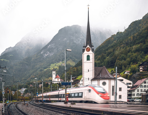 Scenic landscape of Swiss Alps in rainy weather: High-speed train of Swiss railways SBB arriving at Flüelen station (Switzerland), passing church and mountains covered in fog. photo