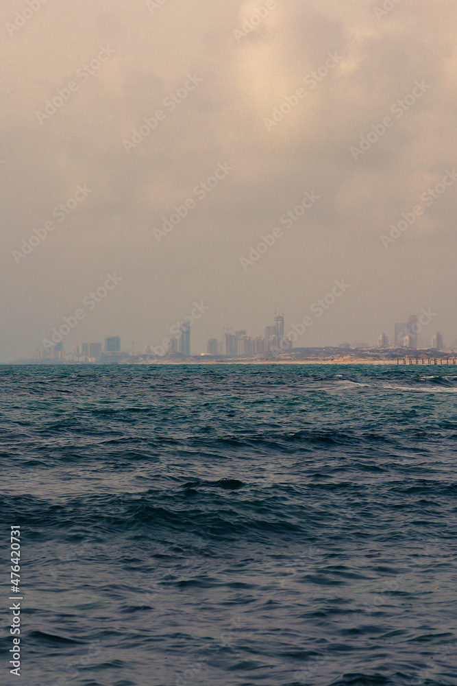 Landscape photo of a cityscape beyond the sea shore. At the front is the blue ocean on a gloomy day.