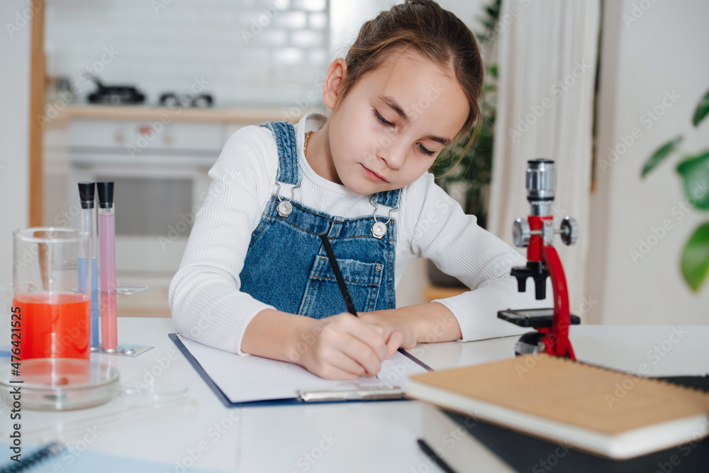 Diligent little girl doing home science project, documenting the process. She has chemistry glassware with colorful liquids and microscope Diligent little girl doing home science project, documenting the process. She has chemistry glassware with colorful liquids and microscope