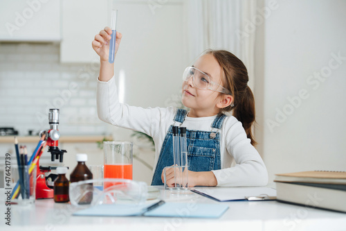 Joyous little girl doing home science project, looking at blue liquid in a flask. She is wearing plastic glasses and has glass chemical equipment and microscope