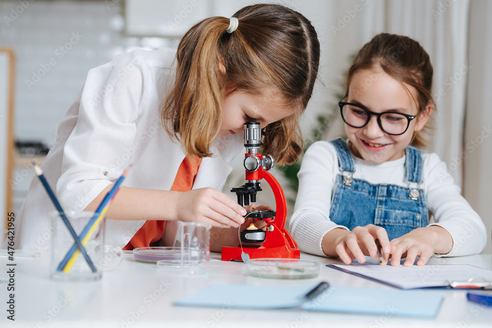 Girl with pony tails looking through the microscope, girl taking notes next to her. Behind a table with books and colored liquids. Girl with pony tails looking through the microscope, girl taking notes next to her. Behind a table with books and colored liquids.