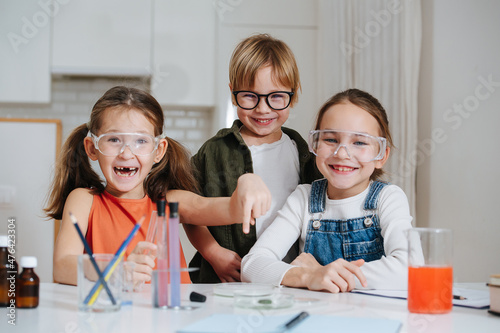 Social little kids doing home science project, sharing their fun experience. All behid table, wearing glasses. Chemical glassware and colored liquids on the table. photo