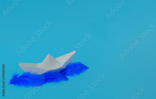 Boat made of white paper on blue feathers, on a sky blue background. Text space. Vacation time, travel, creativity, concept. Minimal style.