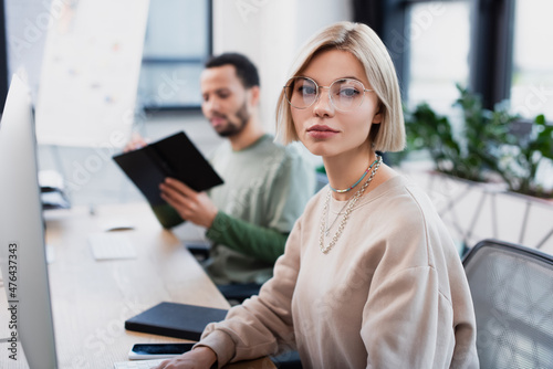 blonde woman in glasses looking at camera near blurred african american colleague.