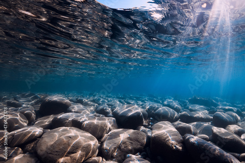 Underwater view with stones bottom, sun rays and reflection in water.