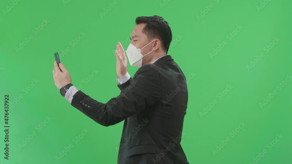 Side View Of Asian Business Man Having Video Call On Mobile Phone While Walking On Green Screen Chroma Key, Wear Medical Mask
