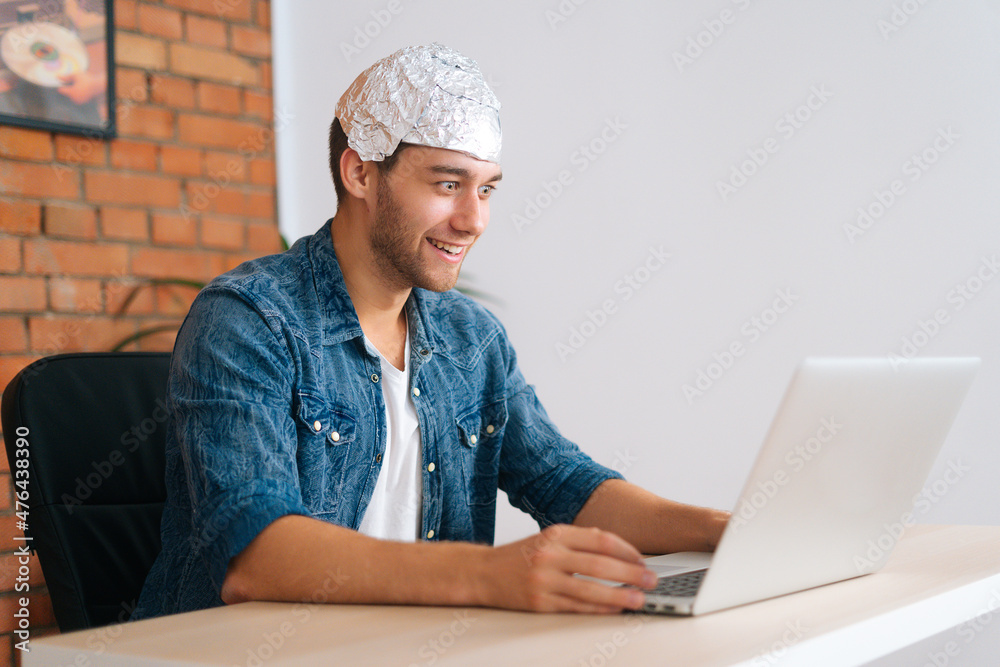 Side view of crazy smiling male conspiracy theorist wearing aluminum foil protect brain watching shocked online video content using laptop computer, sitting at desk in living room with modern interior Side view of crazy smiling male conspiracy theorist wearing aluminum foil protect brain watching shocked online video content using laptop computer, sitting at desk in living room with modern interior