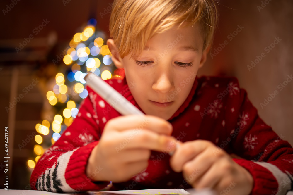 a boy in a red Christmas sweater on the background of a Christmas tree is engaged in the process of drawing in his room under a lamp a boy in a red Christmas sweater on the background of a Christmas tree is engaged in the process of drawing in his room under a lamp