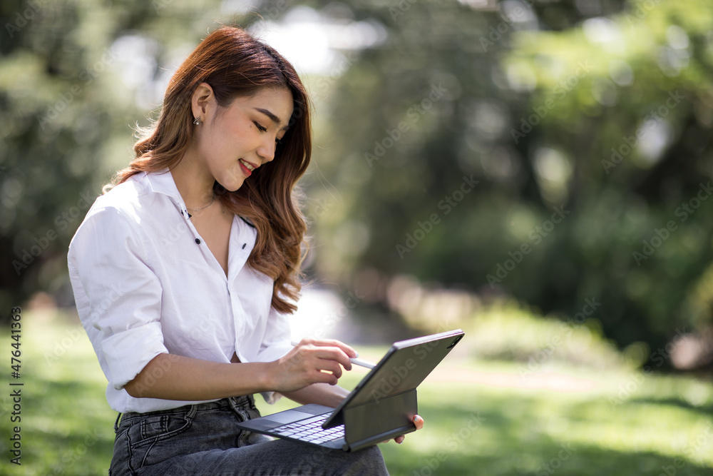 Fototapeta premium Joyful casual Asian student girl with laptop outdoors. Smiling woman sitting on the grass with a computer, surfing the net or preparing for exams. Technology, education and remote working concept