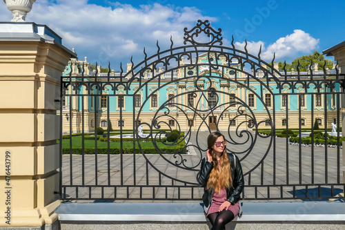 A girl with long hair and in a shot dress sits on a gate leading to a palace. View on the alace is disturbed by the fence. CLear and blue sky. Girl is enjoying her time. photo