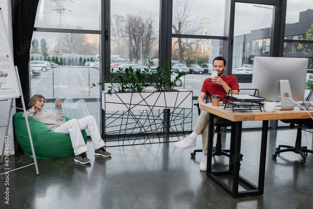 Fototapeta premium blonde woman sitting in bean bag chair and using smartphone near african american colleague in office.