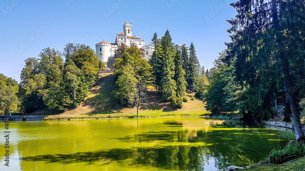 A view on Trakoscan castle from the lake side. Castle is located on a small hill and well hidden in between the trees. The lake in front of the castle is really green. Clear and bright day. A view on Trakoscan castle from the lake side. Castle is located on a small hill and well hidden in between the trees. The lake in front of the castle is really green. Clear and bright day.