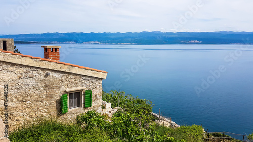 A house made of stones with green shutters located on a hill with a marvellous view on the sea. Each corner of the house is overgrown with small vine and bushes. Calm surface of the sea. Hidden gem photo