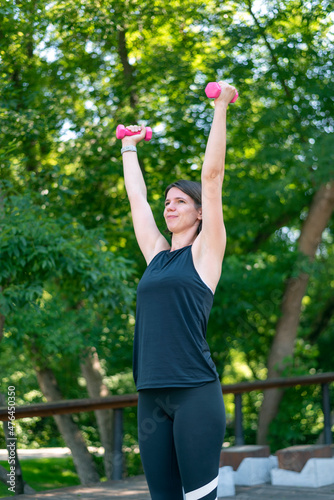Young athletic woman does fitness with dumbbells outdoors. Girl holds the dumbbells with her hands up against the background of green foliage.