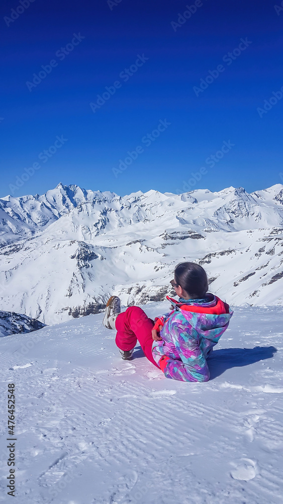 A girl in skiing outfit lies on the snow and enjoys the view on Alps in front of her. The sky is clear blue. Girl is looking front at the tall mountains. Slopes are perfectly graveled