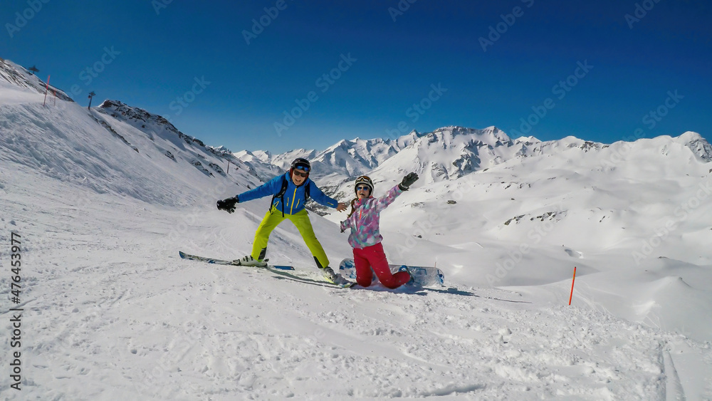 A snowboarder girl and skier boy playing on the slope on a sunny, winter day. Girl is kneeling on the slope. They are happy and excited. Tall alpine mountains behind the couple. Clear and bright day.