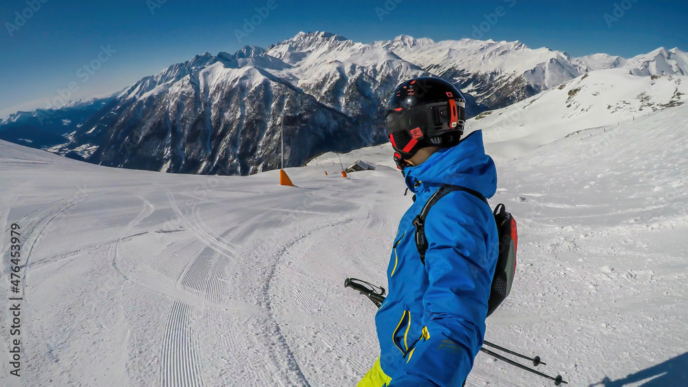 A young skier takes a selfie while skiing down a perfectly groomed slope. Man is wearing helm for the protection. Tall Austrian Alps in the back. Each peak and slope is covered with thick snow layer.