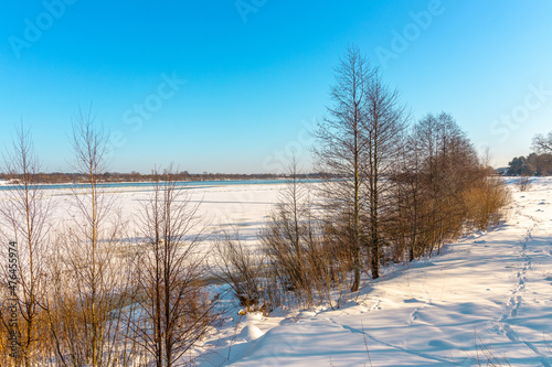 Beautiful winter landscape with river shores, trees, snowy meadow and blue sky in background