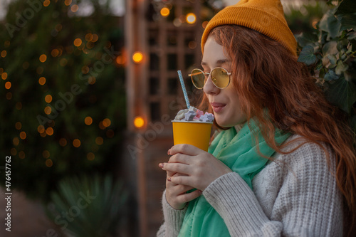 X mas time, red hair girl walking in park on a cold day, enjoying cup of hot chocolate or cocoa, wearing warm in head wear, yellow hat, knitted sweater against the decorated lights