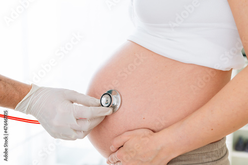 Latex gloved hand of a doctor using a stethoscope on the belly of a pregnant woman in a pregnancy check-up to check the baby's heartbeat before birth.
