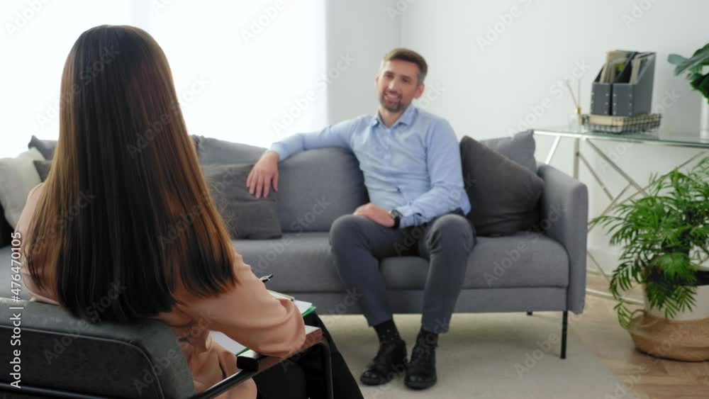 Psychology of mental health. Unfocused smiling adult man patient sitting on couch talking to psychologist at psychotherapy session. Woman psychologist listens client sitting on chair in clinic office Psychology of mental health. Unfocused smiling adult man patient sitting on couch talking to psychologist at psychotherapy session. Woman psychologist listens client sitting on chair in clinic office