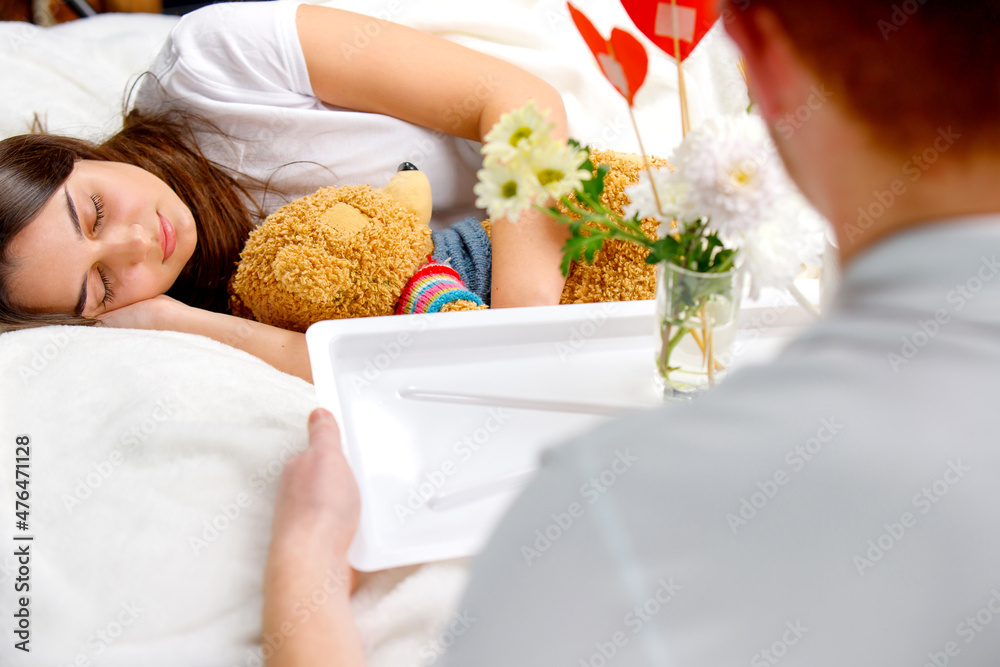 A man holding the tray with a vase of flowers and red heart shape from cardboard to make romantic breakfast for his girlfriend sleeping hugging a bear toy.