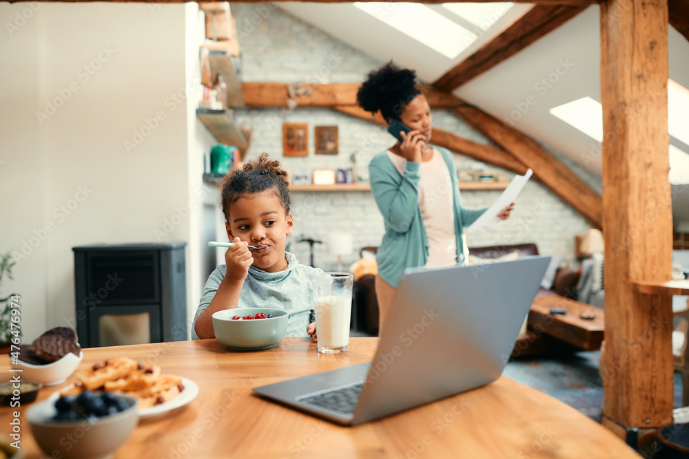 Small African American girl eats breakfast while using laptop at dining table. Her mother in on the phone in the background.