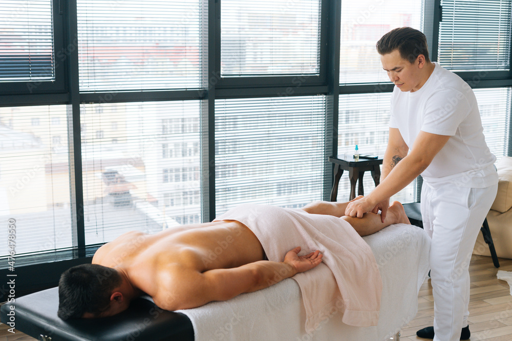 Professional male masseur with strong tattooed hands massaging lower leg of muscular athlete man after physical sports workout, lying on stomach at massage table on background of window