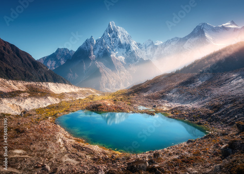 Snowy mountain with illuminated peaks and small lake with blue water at sunrise in Nepal. Beautiful landscape with lake, high rocks in snow, golden sun rays, hills at dawn. Himalayan mountains. Nature