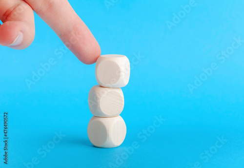 Finger knocking over a small tower of three blank wooden cubes, blocks, pushing and destabilizing a built structure, loss of balance simple abstract concept. Object closeup, detail, blue background photo