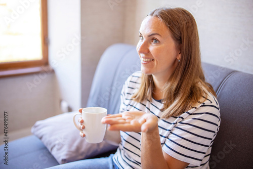 Young excited woman with coffee cup sitting on a sofa near the window talking. Woman sharing latest news or rumor with friend or colleague at home