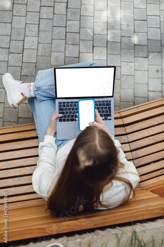 Top view, close-up of smartphone with blank screen in hands of young woman sitting at wooden bench and touching screen. Laptop with blank screen. Mock up space for ad.