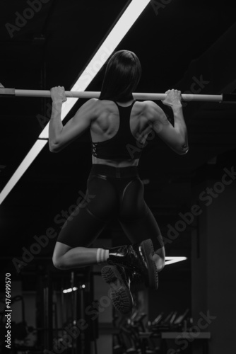 A photo from behind of the sporty woman who is doing wide-grip pull-ups in a gym. A muscular brunette girl wears a black top and high waist stretchy shorts in the back workout.