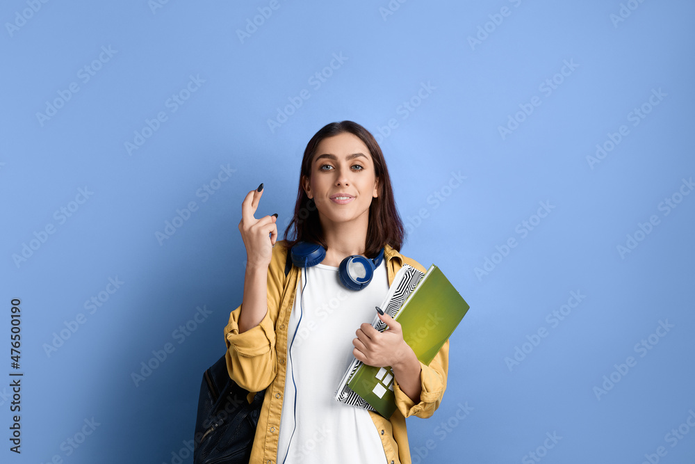 Positive smiling student girl is holding fingers crossed for passing an important exam, hopes for good results, holding books, wearing yellow shirt, white t-shirt, black bag and headphones over neck. Positive smiling student girl is holding fingers crossed for passing an important exam, hopes for good results, holding books, wearing yellow shirt, white t-shirt, black bag and headphones over neck.