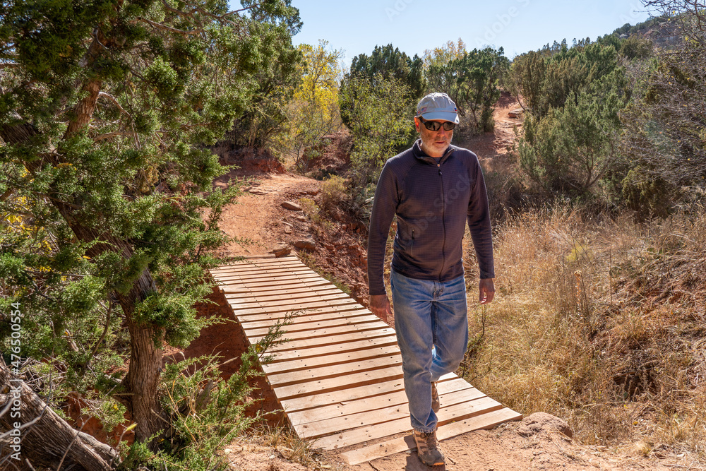 Mature Caucasian man walking across a dry stream on a wooden plank bridge along a dirt trai, frontl, Kiowa Trail, Palo Duro State Park, Texas