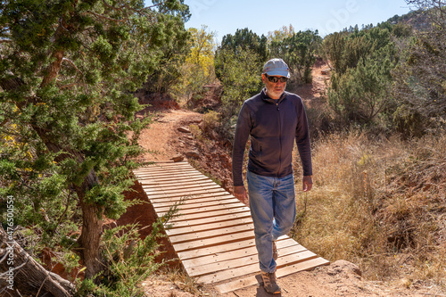 Mature Caucasian man walking across a dry stream on a wooden plank bridge along a dirt trai, frontl, Kiowa Trail, Palo Duro State Park, Texas photo