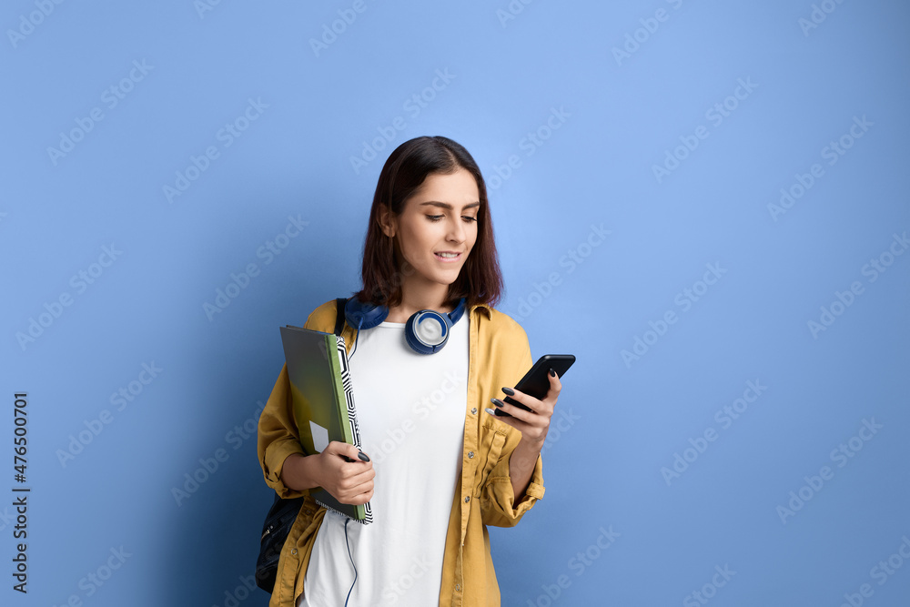 Worried student girl is holding mobile phone in hand, looks at screen and bites lip, scared of checking results, holding books, wearing yellow shirt, white t-shirt, black bag and headphones over neck. Worried student girl is holding mobile phone in hand, looks at screen and bites lip, scared of checking results, holding books, wearing yellow shirt, white t-shirt, black bag and headphones over neck.