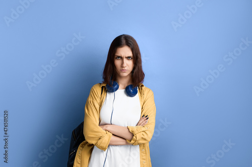 Serious displeased student girl is frowning and crossing arms over chest, does not believe promises, suspects something bad, wearing yellow shirt, white t-shirt, black bag and headphones over neck. © 5M