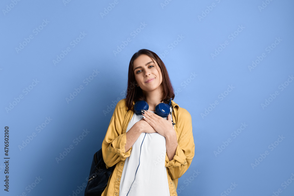Romantic happy student girl is pressing hands to heart, is demonstrating her feelings, in love with brand, service or product, wearing yellow shirt, white t-shirt, black bag and headphones over neck. Romantic happy student girl is pressing hands to heart, is demonstrating her feelings, in love with brand, service or product, wearing yellow shirt, white t-shirt, black bag and headphones over neck.