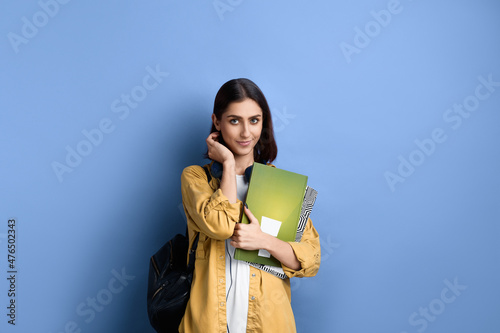 Timid pretty student girl is tucking hair behind the ear, smiles, met boyfriend, feels she is looking adorable, holding books, wearing yellow shirt, white t-shirt, black bag and headphones over neck. photo