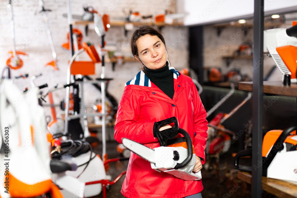 European woman standing in salesroom of gardening tools store with chainsaw in hands and looking in camera.