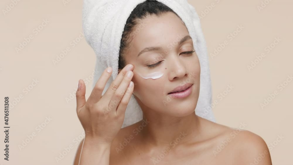 Good-looking African American young slim woman with a towel on her head and nude shoulders puts white cream on her cheek and smiles wide for the camera on beige background | Skincare cream commercial Good-looking African American young slim woman with a towel on her head and nude shoulders puts white cream on her cheek and smiles wide for the camera on beige background | Skincare cream commercial