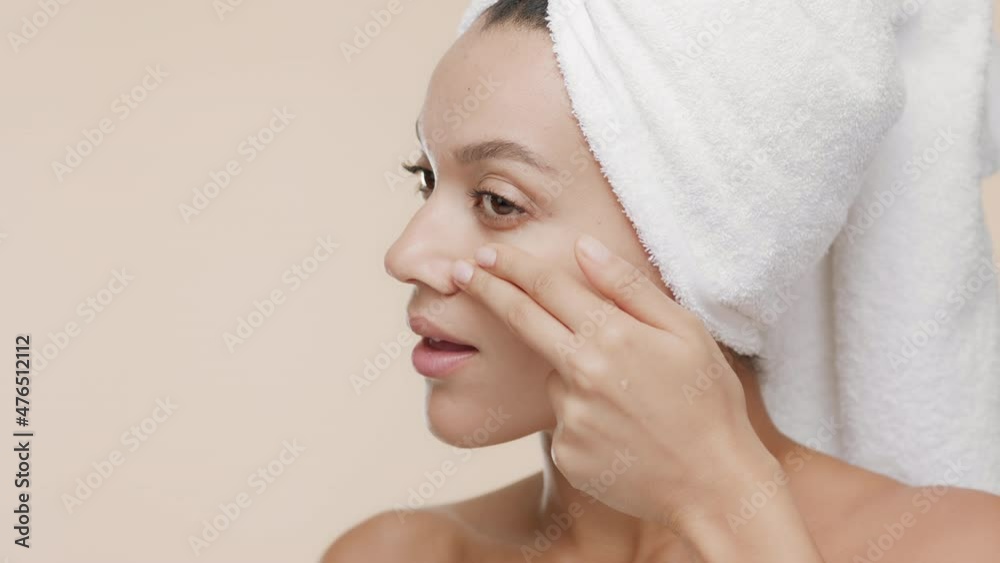 Cute African American young slim woman with a towel on her hair and bare shoulders applies white cream on her cheek and smiles wide for the camera on beige background | Moisturizer commercial Cute African American young slim woman with a towel on her hair and bare shoulders applies white cream on her cheek and smiles wide for the camera on beige background | Moisturizer commercial