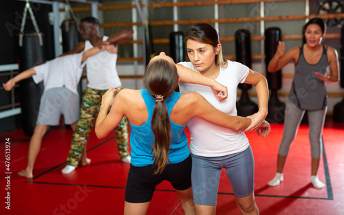 Latin woman training elbow strike with another woman during group self-protection training. Asian woman instructor standing nearby and correcting them.