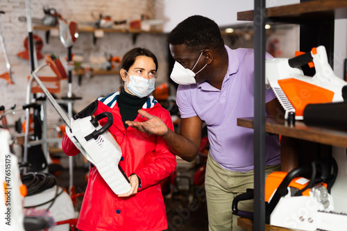 African-american man in face mask consulting Caucasian woman about chainsaw in gardening tools store.